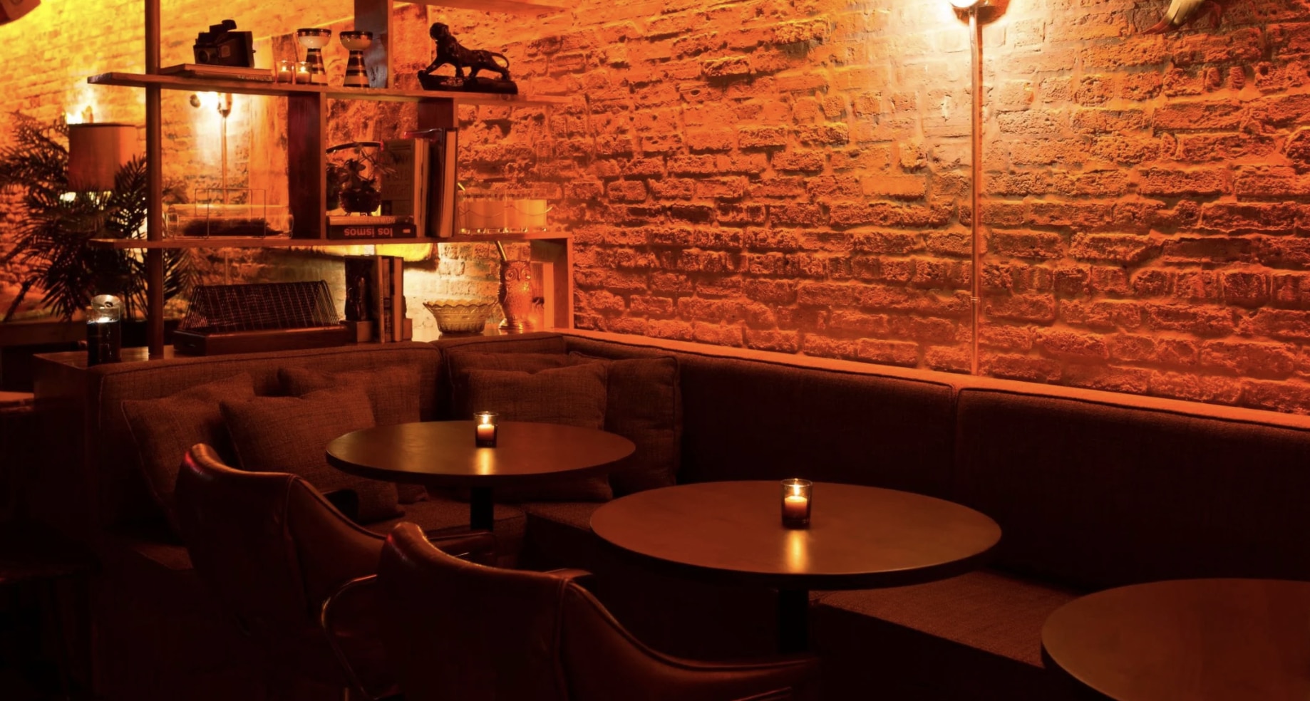 chicago speakeasy bar that is behind a barber shop, and has circle tables with a candle and a brick wall in the back with red and yellow lighting