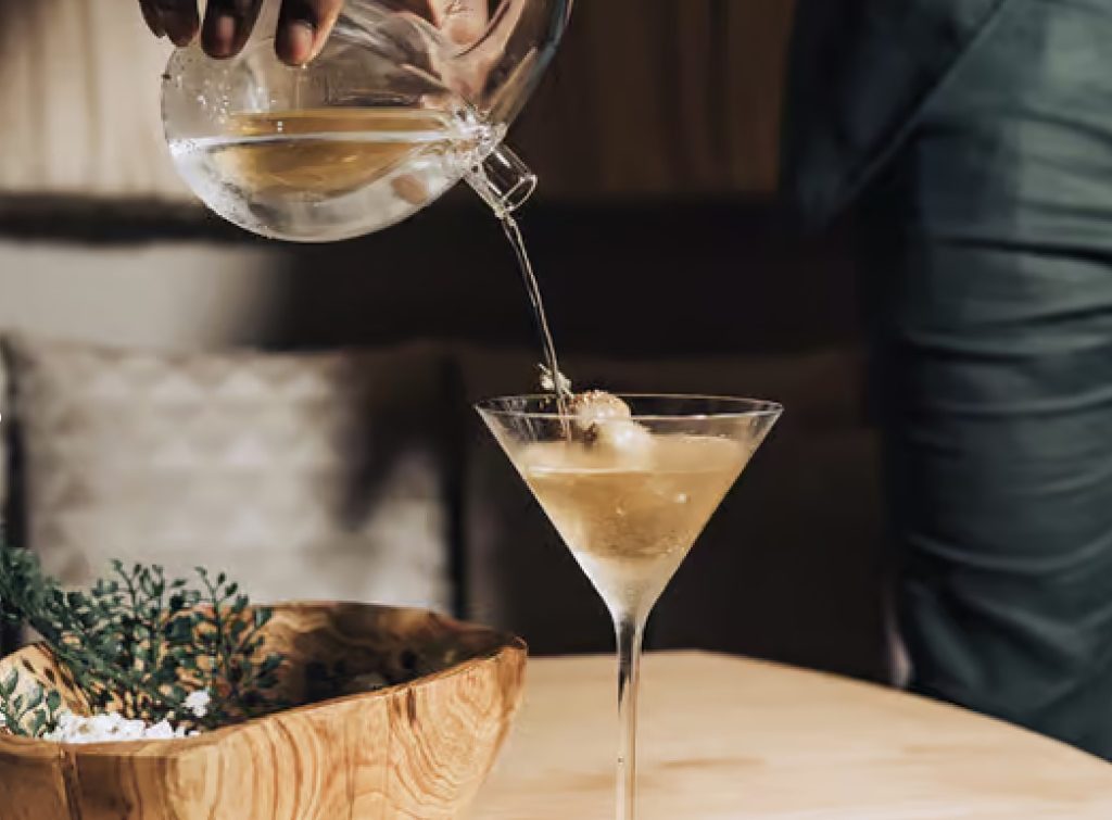 someone pouring a cocktail in a martini glass at nisos on a wooden table