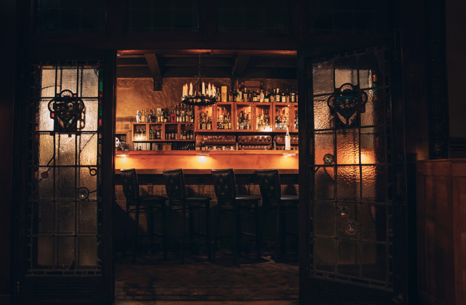 dimly lit bar with 4 stools, candles, and a collection of spirits in the back of the bar 