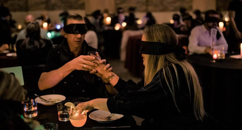 couple blindfolded at a dinner table with clinking their drinks and trying food