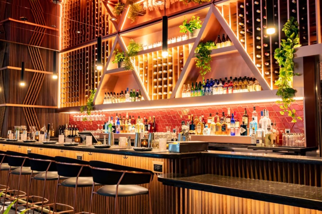 the bar with top shelf bottles at planta queen with a neon light up triangle sign backsplash and stools for seating