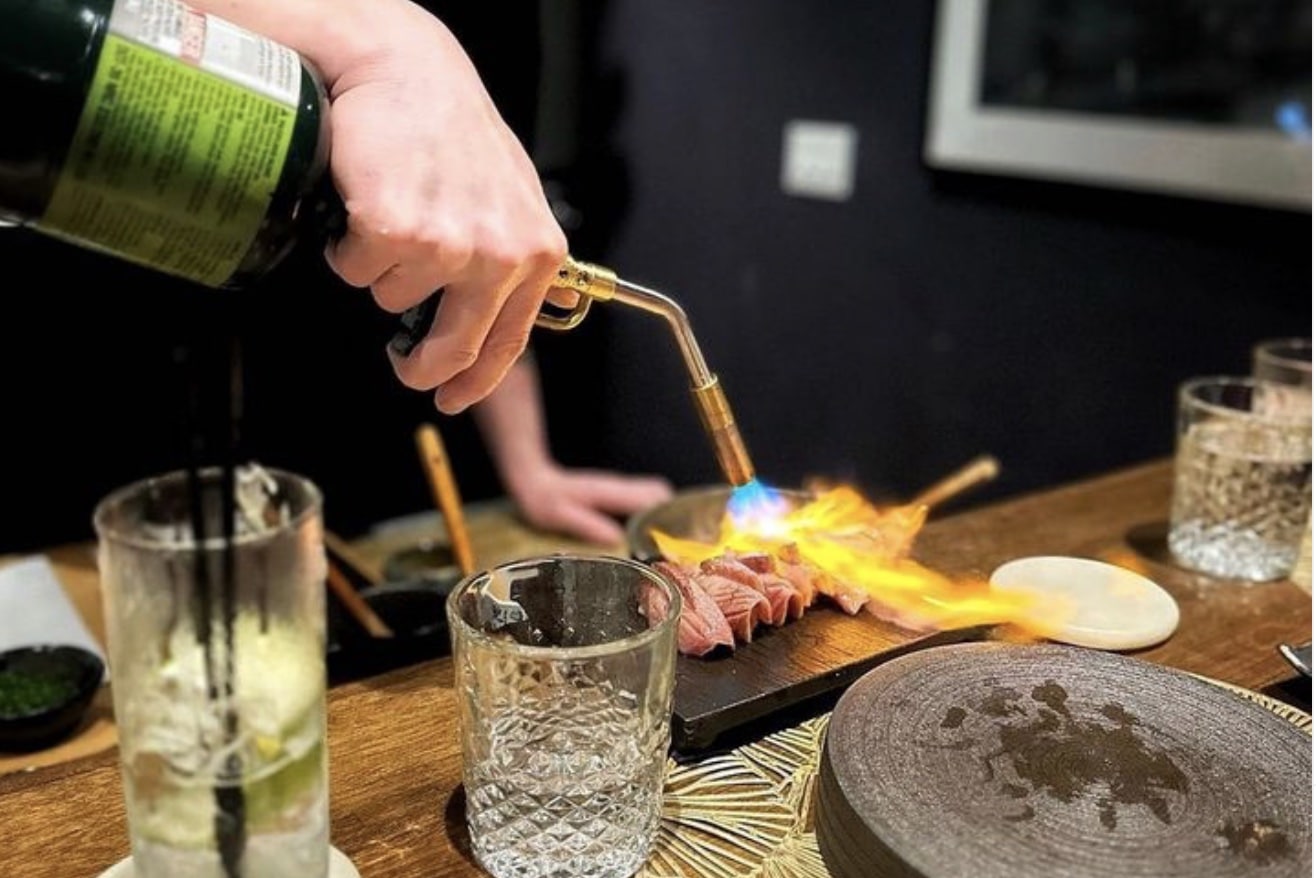 someone using a flame seared fish with drinks next to the plate from sushi suite in chicago