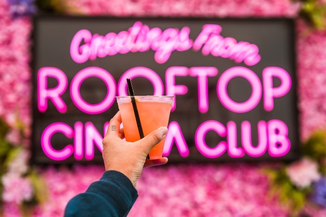 Person holding a drink in front of the Rooftop Cinema Club neon sign 