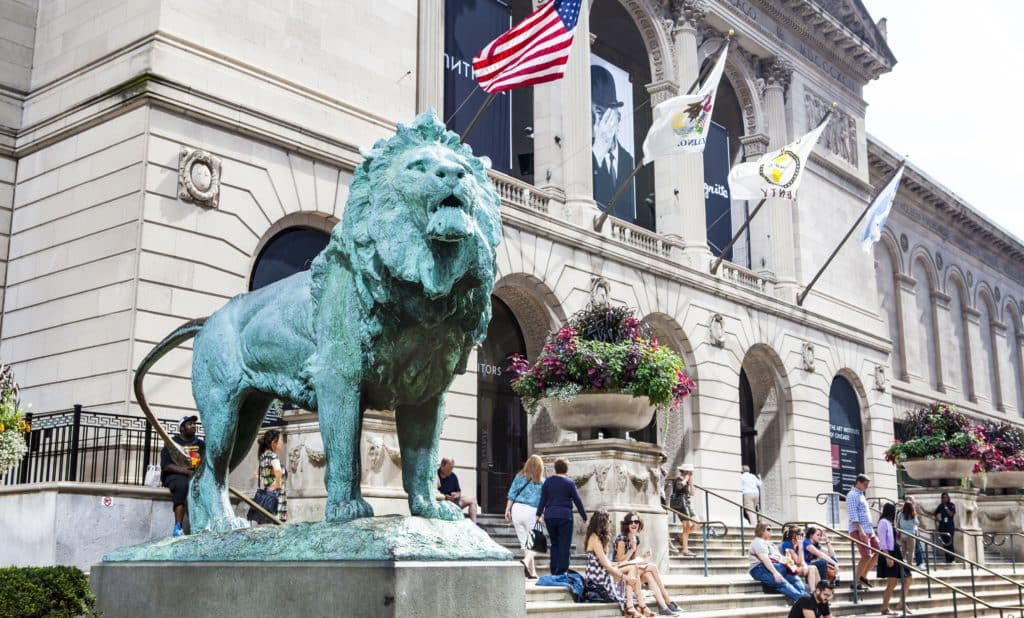 the American flag behind a large stone lion outside the art institute of Chicago with people on the steps