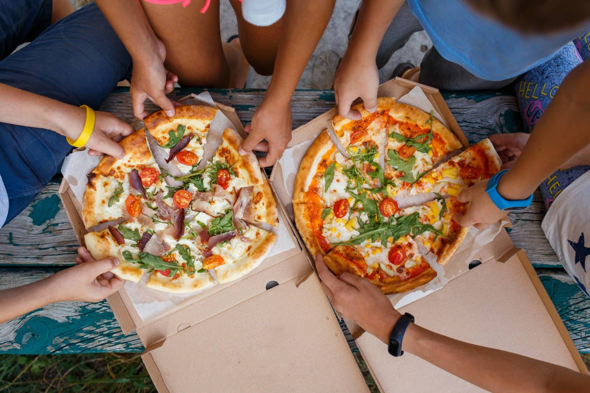 hands grabbing for slices of two large pizzas in cardboard boxes 