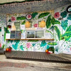 Front facade of the donut shop shows walls painted with colorful tropical leaves and flowers along with an open window for ordering