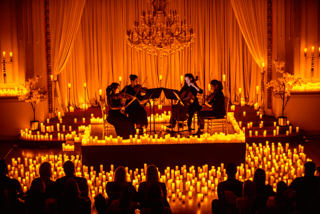 A string quartet performs on a raised stage, surrounded by hundreds of candles, with a silhouette of audience members watching