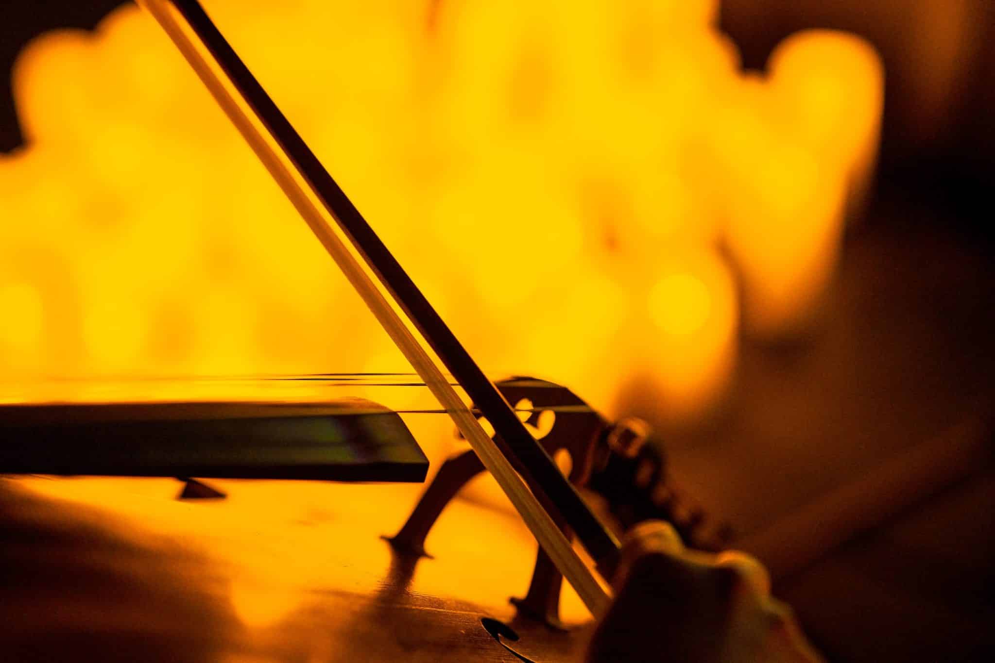 A close-up of a bow being used to play a string instrument with the glow of candles in the background at a Candlelight concert.