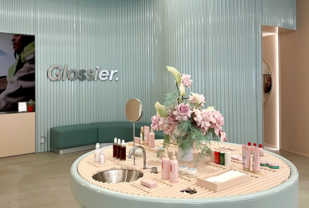 Glossier Chicago showroom with blue wall, silver logo and products displayed on a white round table