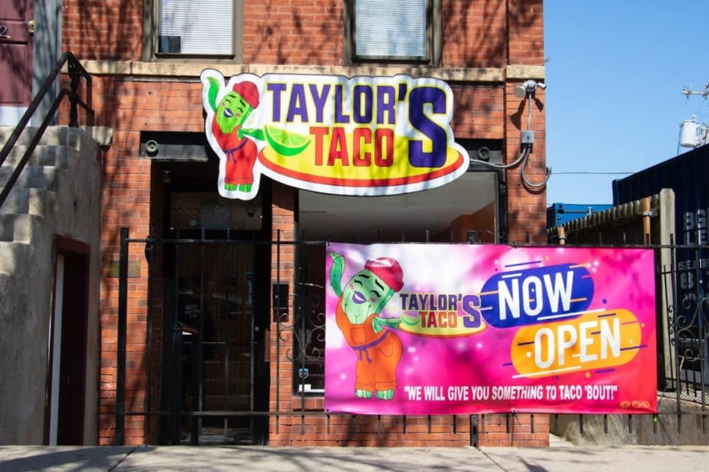 Exterior of Taylor's Tacos in Little Italy with a bright name tag sign and a opening banner