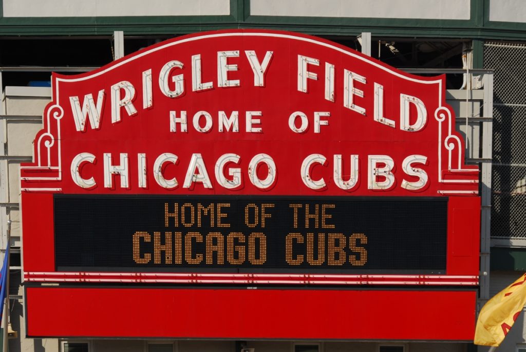 The classic Wrigley Field sign says 'Wrigley Field Home of the Chicago Cubs' in big lettering with a message board style box reading 'Home of the Chicago Cubs'