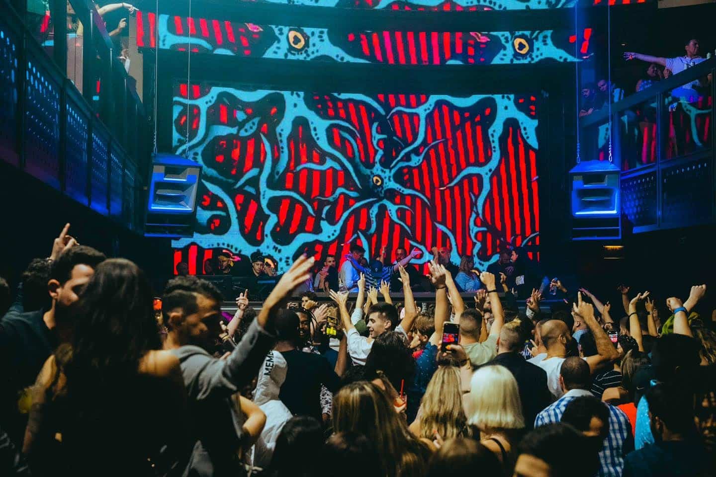 people with their hands in the air, dancing to the tunes of the dj at prysm with a electronic painting behind the dj