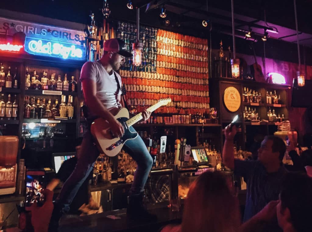 man in a hat playing the guitar on top of the bar with people waving their ehands below at bub city in Chicago