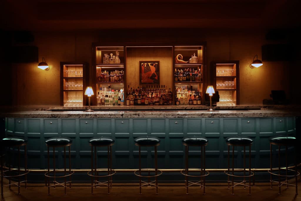 interior of lazy bird with a blue bar counter and stool in front of a a stocked bar with alcohol bottles and dim lights in fulton market chicago