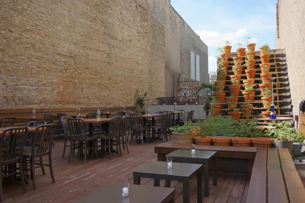 brick wall with wooden tables and chairs with candles unlit and a stack of potted plants behind at taxim rooftop in Chicago