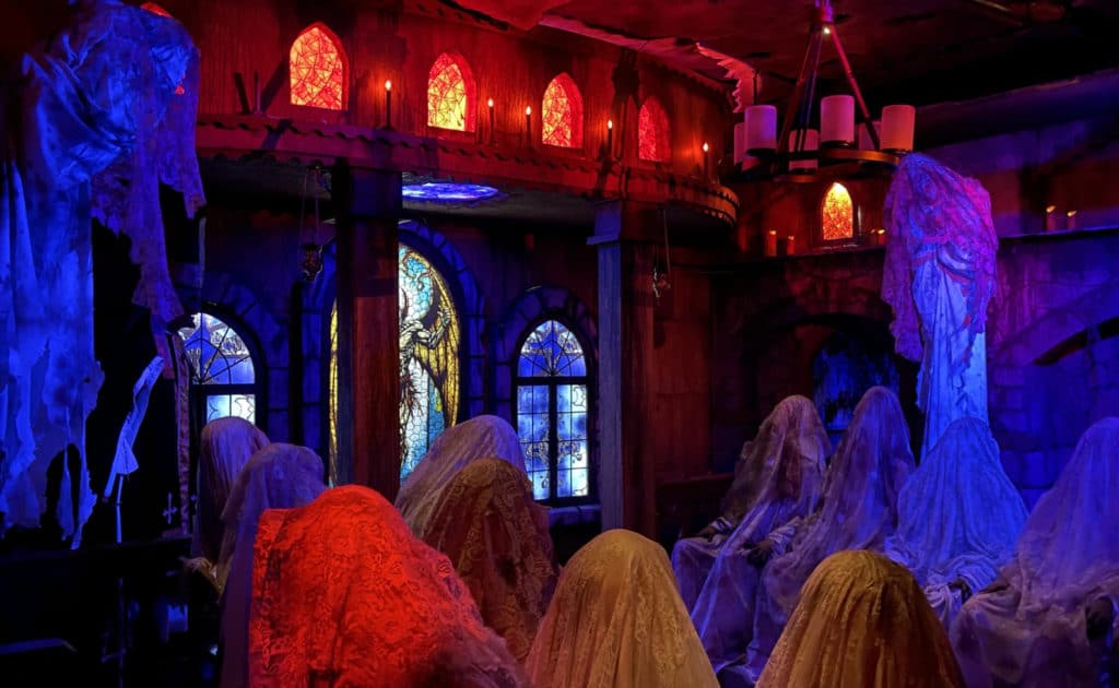 a room filled of ghosts underneath candlelight with red and blue lights shining through and a stained glass window in the back at Dungeon of Doom Haunted House