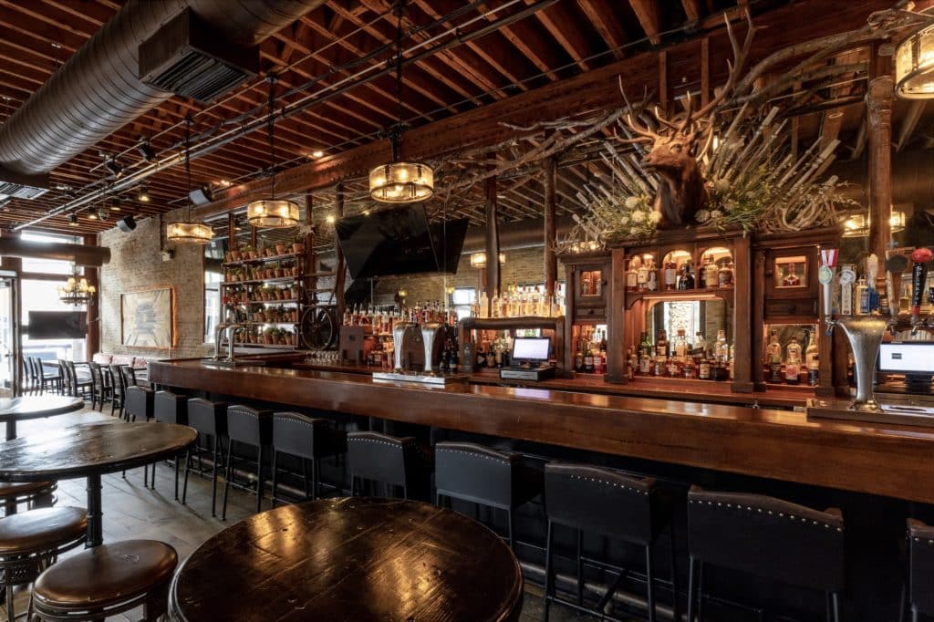 interior of frontier bar with wooden beams, wooden tables, and a wooden bar with chairs and a bar in the background with vodka, gin, whiskey, and plants