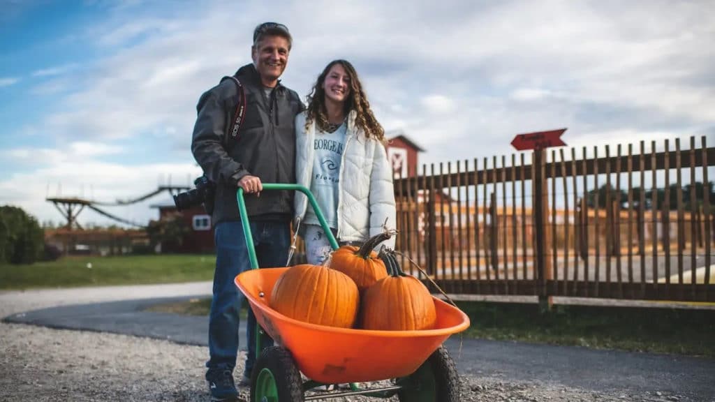 father and daughter holding an orange barrel with three pumpkins and a gate in the background