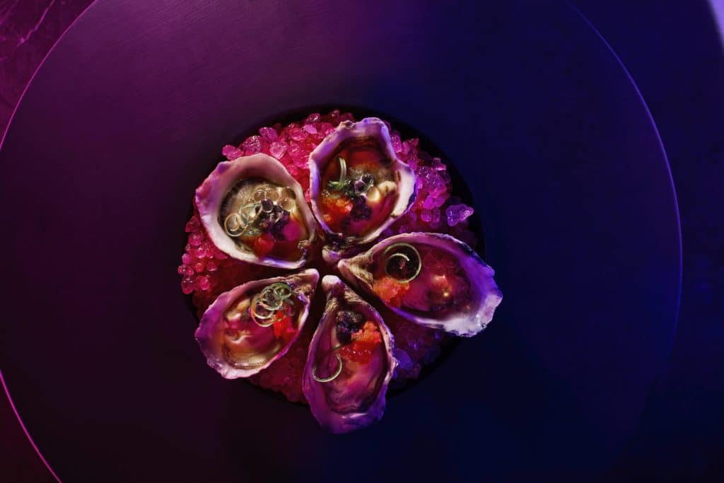 five oysters with garnishes on ice with purple light in the background