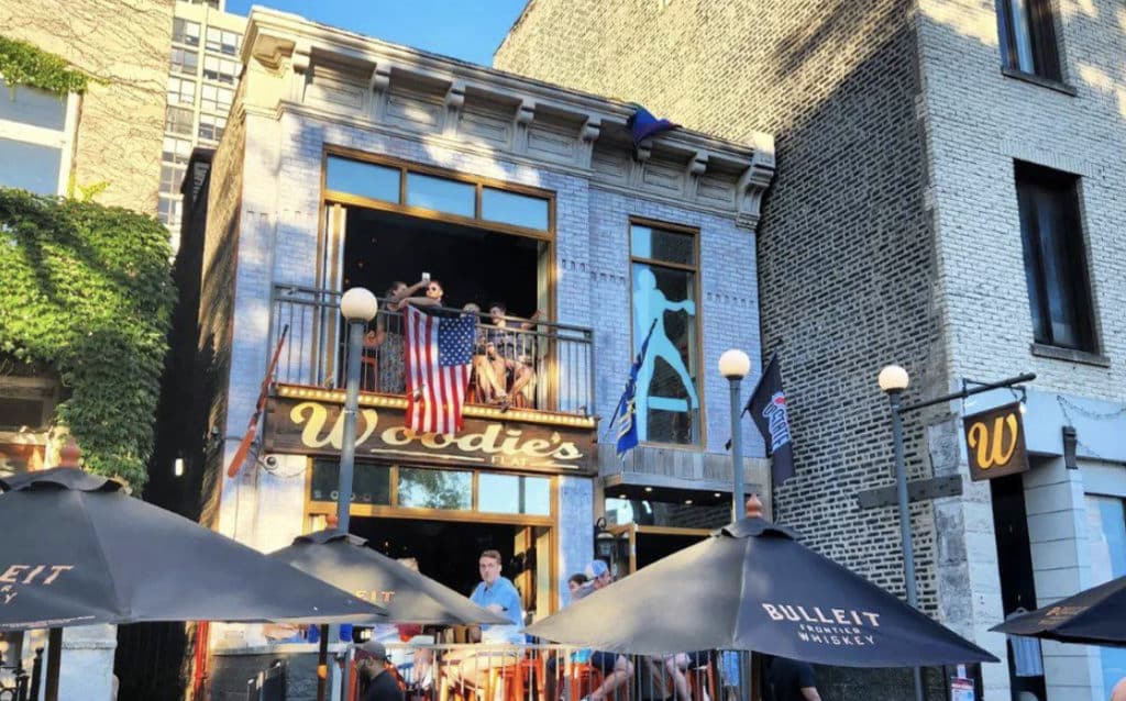 stone exterior of woodie's flat with an american flag hanging out the window and people on the second floor drinking with umbrellas on the patio
